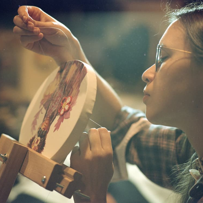Winona Nelson, an Indigenous artist, carefully stitches beadwork onto an embroidery hoop while holding a needle and thread. Wearing glasses and concentrating on the piece, the artist works on a floral design that combines painted imagery with intricate beadwork, highlighting the precision and craftsmanship of the process.