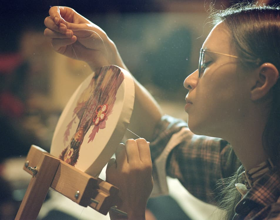 Winona Nelson, an Indigenous artist, carefully stitches beadwork onto an embroidery hoop while holding a needle and thread. Wearing glasses and concentrating on the piece, the artist works on a floral design that combines painted imagery with intricate beadwork, highlighting the precision and craftsmanship of the process.