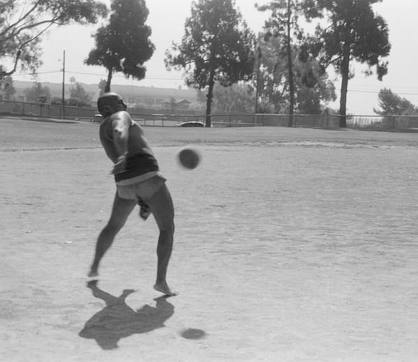 Indigenous Ulama player known as “Nu” swings his body to strike a rubber ball with his hip during a match on an outdoor dirt field. The fast-moving ball is captured midair as trees and a fence line the background.