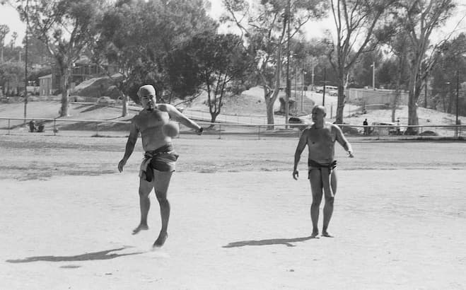 Two Indigenous players compete in a game of Ulama on a dirt field. Captain Marco "Kenwel" Santana, one player in the field, leaps into the air as he strikes the heavy rubber ball with his hip, while another player watches from behind. Both wear traditional loincloths, and trees and park buildings are visible in the background.