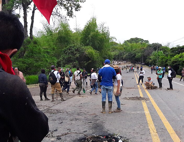 Indigenous community members participate in a protest in Cauca, Colombia. People gather on a rural roadway, some standing across the street while others observe nearby. The demonstration was led by Indigenous communities, primarily Nasa people, with representation from the CRIC movement.