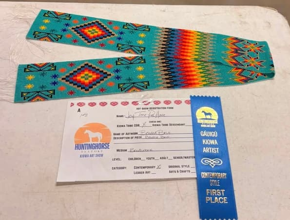 A turquoise beaded belt featuring colorful geometric Kiowa-inspired designs and a rainbow-striped center is displayed on a white table. Below it are the artist registration form and a blue First Place ribbon from the Hunting Horse Kiowa Art Show, recognizing the belt in the Contemporary Style category.
