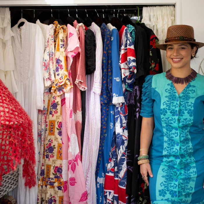 A woman stands smiling to the right of her open closet, displaying a selection of colorful vintage dresses. She is wearing a two-tone turquoise Muʻumuʻu with darker blue flowers printed on a light blue base. The dress has puffed sleeves and buttons down the front. She is wearing a necklace made of purple sea urchin spikes, a woven brown hat with a brown and blue feathered lei wrapped around the brim, and a mix of gold, jade, pearl, and lauhala bracelets down her arms. Her hair is pinned up, and she is wearing button earrings made of Niʻihau shells. There is a lamp on the other side of the closet covered in a bright pinkish-red crochet. A circular mirror hangs behind, with hooks under it, displaying her necklace collection.