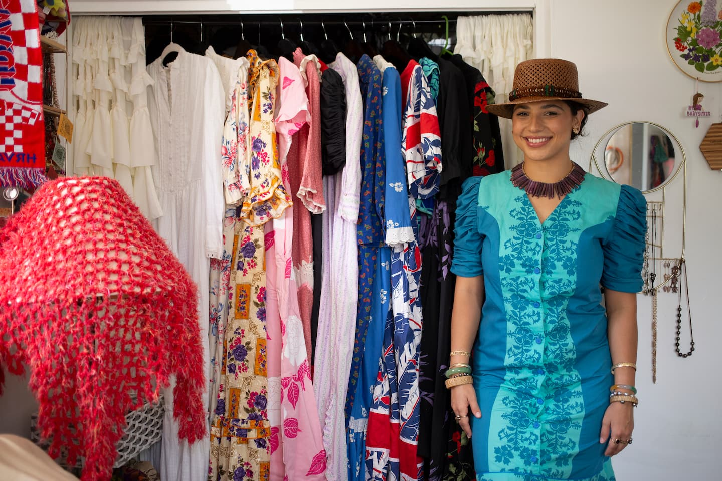 A woman stands smiling to the right of her open closet, displaying a selection of colorful vintage dresses. She is wearing a two-tone turquoise Muʻumuʻu with darker blue flowers printed on a light blue base. The dress has puffed sleeves and buttons down the front. She is wearing a necklace made of purple sea urchin spikes, a woven brown hat with a brown and blue feathered lei wrapped around the brim, and a mix of gold, jade, pearl, and lauhala bracelets down her arms. Her hair is pinned up, and she is wearing button earrings made of Niʻihau shells. There is a lamp on the other side of the closet covered in a bright pinkish-red crochet. A circular mirror hangs behind, with hooks under it, displaying her necklace collection.