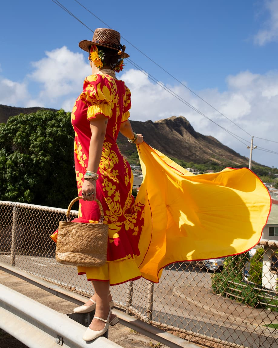 A woman stands atop a street barrier railing, looking out at the street below and off into the distance at a pointed mountain peak. The sky is bright blue, and there are clouds off on the horizon. She is wearing a red vintage dress with yellow flowers and a yellow bustle detached from the back of the dress, held in her right hand and billowing in the wind. In her left hand, she holds a square brown woven purse with curved bamboo handles. She wears a dark brown woven hat and two yellow-and-red floral hair clips on either side of her bun.