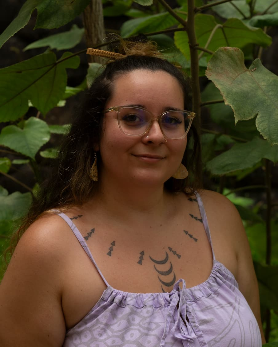 A head and shoulders portrait of a woman standing in front of large heart-shaped dark green leaves. She is wearing a light purple spaghetti-strap dress that cinches at the front and ties into a bow. The pattern on the dress is printed in light grey and consists of hexagonal shapes with dots in their centers. She has a tattoo of crescent-moon shapes and columns of small triangles lining her chest in a curved pattern from shoulder to shoulder. She is wearing her hair in a half-up, half-down style, with a bun at the top of her head, held in place by a bamboo hair pick carved with a pattern on one end. She is wearing clear glasses with three crystals on the sides. She has a nose piercing in either nostril and is wearing earrings made of a round paper-like material printed with a black inked pattern.