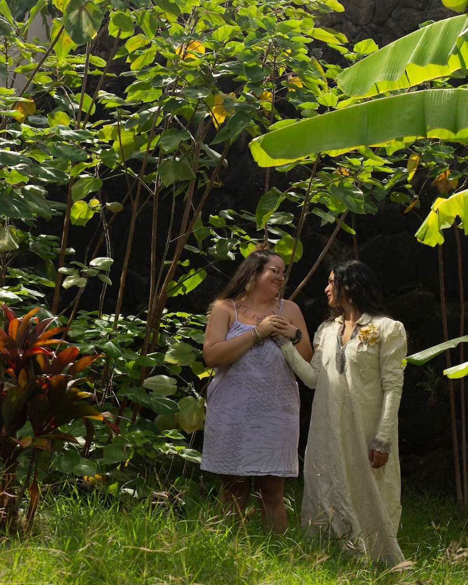 Two women stand side by side, looking lovingly toward one another. The woman on the right has long dark hair and is wearing a vintage-style dress made of a white bark cloth. The woman on the left has long brown hair, half-up in a bun. She is wearing a light purple spaghetti-strapped dress and is holding the other woman's hand to her chest in both hands. They stand together within a tropical garden shaded by the heart-shaped leaves of tall mulberry trees.