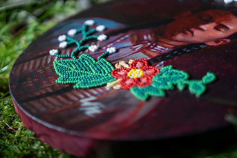 A close-up of intricate floral beadwork in green, red, yellow, and white is stitched onto a painted circular artwork featuring the portrait of an Indigenous person. The detailed beadwork overlays the painted surface, blending traditional bead artistry with contemporary visual art.