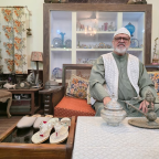 An elderly man sits in the living room of his home, dressed in traditional attire and surrounded by family heirlooms and decorative objects. On a wooden table in front of him are two pairs of handcrafted silver footwear—a pair of silver nagras and a pair of ornate silver wedding sandals—highlighting the enduring cultural significance of these traditional crafts. A glass display cabinet filled with ceramics and antiques stands behind him, while patterned cushions and floral curtains frame the warmly decorated room.