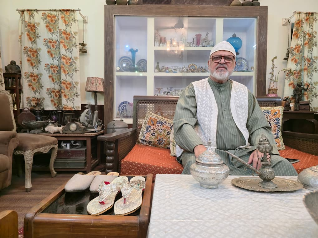 An elderly man sits in the living room of his home, dressed in traditional attire and surrounded by family heirlooms and decorative objects. On a wooden table in front of him are two pairs of handcrafted silver footwear—a pair of silver nagras and a pair of ornate silver wedding sandals—highlighting the enduring cultural significance of these traditional crafts. A glass display cabinet filled with ceramics and antiques stands behind him, while patterned cushions and floral curtains frame the warmly decorated room.