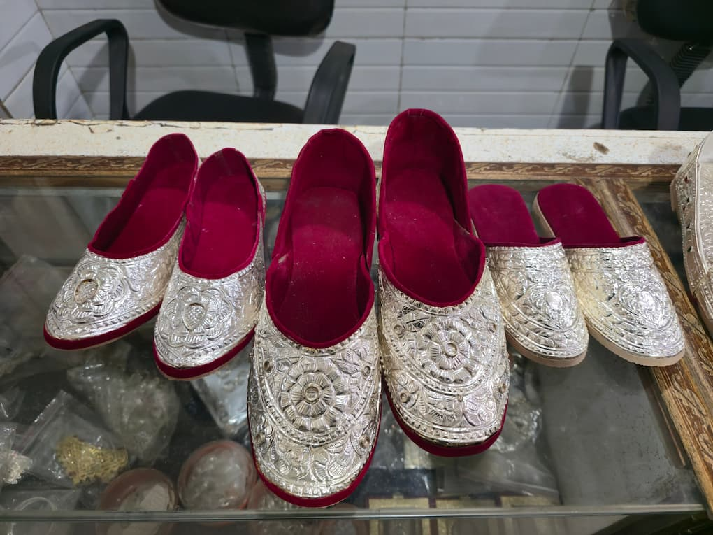 Three pairs of handcrafted silver nagras with deep red velvet lining are displayed on a glass countertop inside a shop. Each pair features intricate silver floral and geometric embossing, reflecting the detailed craftsmanship that has traditionally made the footwear a prized part of wedding attire.