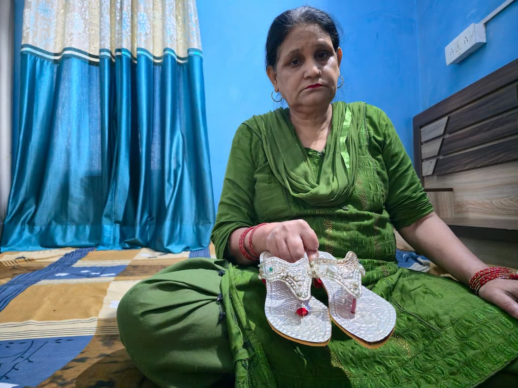 A woman sits on the floor of her home holding a pair of ornate silver wedding sandals in her lap. Dressed in a green outfit, she looks toward the camera while displaying the handcrafted footwear, which features intricate silver embossing. Blue curtains and a wooden bed frame are visible in the background.