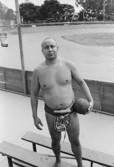 Indigenous Ulama player known as “Nu” stands barefoot on the bleachers beside a playing field, holding a heavy rubber ball. He wears traditional playing attire, including a protective hip wrap and loincloth, and looks directly at the camera as a chain-link fence and the Ulama field stretch out behind him.