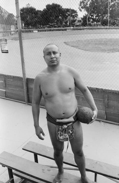 Indigenous Ulama player known as “Nu” stands barefoot on the bleachers beside a playing field, holding a heavy rubber ball. He wears traditional playing attire, including a protective hip wrap and loincloth, and looks directly at the camera as a chain-link fence and the Ulama field stretch out behind him.