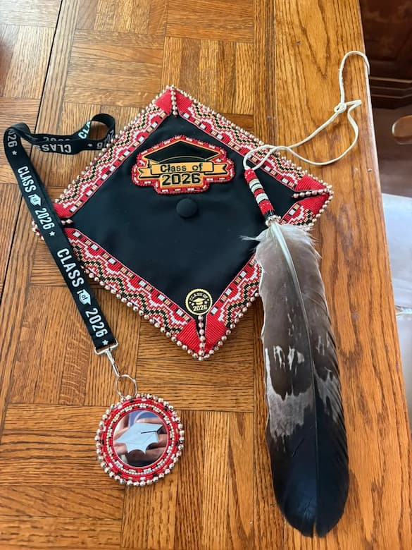 Hand-beaded graduation cap decorated with red, white, black, and gold Indigenous-inspired beadwork around the edges. A beaded "Class of 2026" centerpiece sits near the top, and a matching beaded medallion hangs from a black "Class of 2026" lanyard. A tassel wrapped in coordinating beadwork is adorned with a large feather. The cap is displayed on a wooden floor.