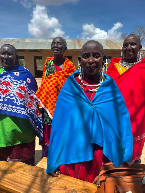 Maasai women stand together during Enkipukonoto Eaji, a traditional naming ceremony for a newborn child. They wear brightly colored traditional clothing and beaded jewelry and smile in front of a village building under a blue sky.