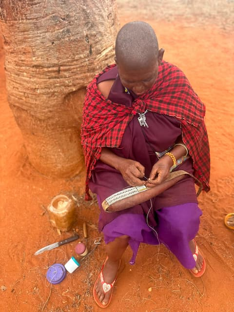 An Elder Maasai woman sits outdoors on the red earth of Northern Tanzania creating traditional Maasai beadwork called Engoti. She wears traditional Maasai clothing while carefully threading beads by hand. Small containers of beads and tools are placed beside her.