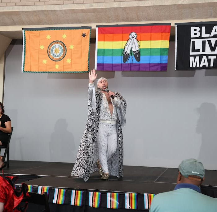 Gay and Two-Spirit artist Tony Enos, a member of the Echota Cherokee Tribe, performs on the DBIWP 2025 stage wearing a white outfit and patterned cape. Behind him are the Cherokee Nation, Indigenous Pride, and “Black Lives Matter” flags. Photo: Washington Talent.