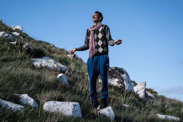 Māori filmmaker Taratoa Stappard stands on a rocky hillside under a clear blue sky, looking upward with a joyful expression.