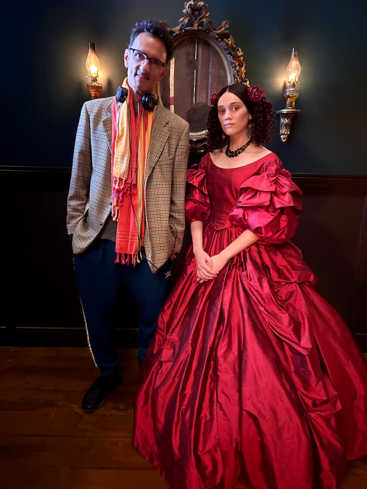 Māori filmmaker Taratoa Stappard and Māori actress Ariāna Osborne stand side-by-side on the set of the film Mārama. They are posing in front of a dark teal wall decorated with an ornate antique mirror and two glowing, warm-toned wall sconces.