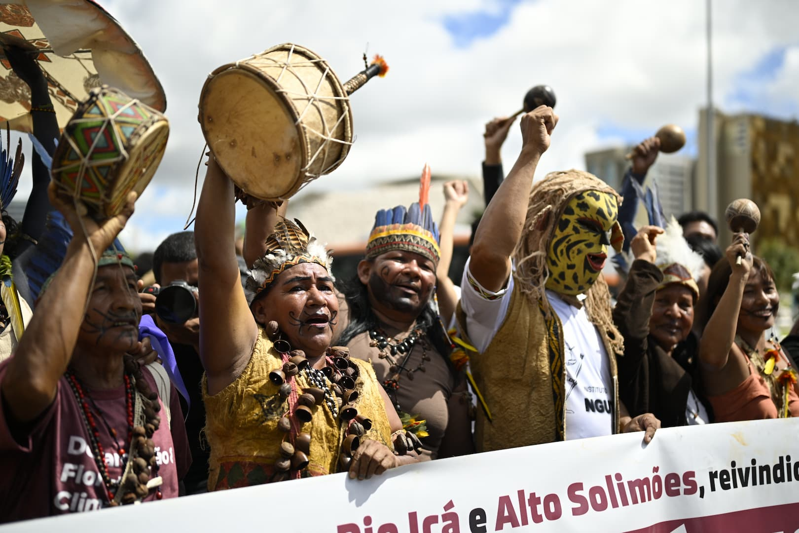 A group of Indigenous people from the Tikuna tribe marches in Brasilía, under a cloudy sky. In the foreground, several members in traditional headdresses and facial markings carry a white banner.