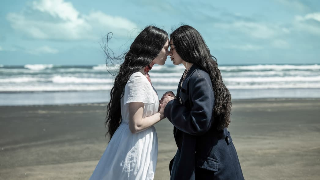 Two Māori women with long, dark hair stand close together on a black sand beach, with their foreheads gently touching. They are holding hands. In the background, white waves crash against the ocean under a pale, overcast sky.