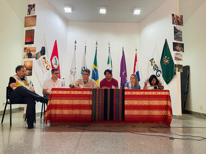 Panelists seated at a table draped in red and orange woven textiles during a Sacred Path discussion, with international flags displayed behind them.