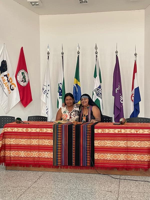 Indigenous women and panelists Kunhã Takuaju and Jovina Renhga sit side by side behind a table covered in a red, yellow, and black traditional textile at the seminar “The Sacred Path of Peabiru: From the Atlantic to the Pacific and Contemporary Integrations in Latin America.” Behind them, a row of institutional, national, and state flags stands against a white wall.