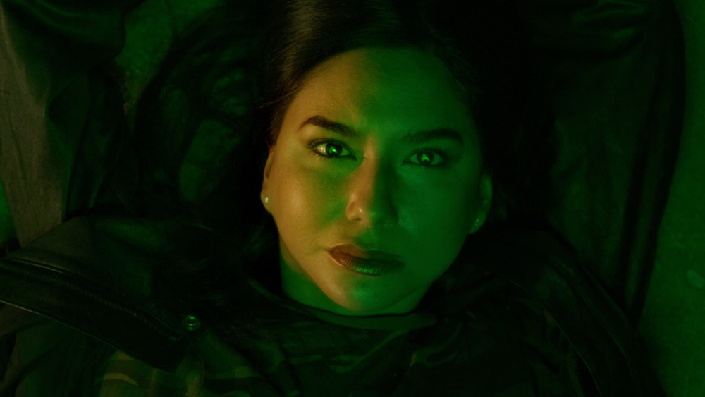 A close-up of an Indigenous woman’s face with long, dark hair, lying down and looking directly at the camera under green and dark lighting. Photo credit: Kwêskosîw: She Whistles (2021)